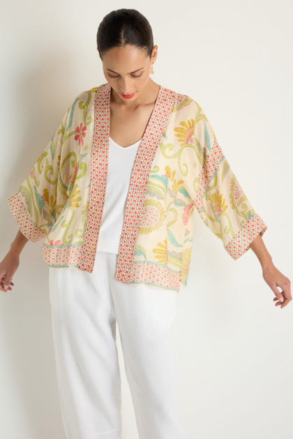 A woman models the Kantha Lime Kimono, featuring a lime-ecru palette and red trim in lightweight woven cloth, styled over a white top and pants against a plain background.