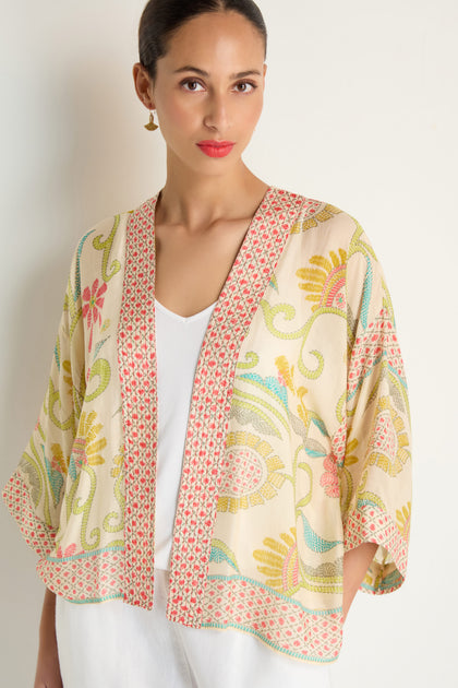 A woman wears the Kantha Lime Kimono, made from lightweight woven cloth with floral lime ecru designs, over a white top and pants against a plain light background.