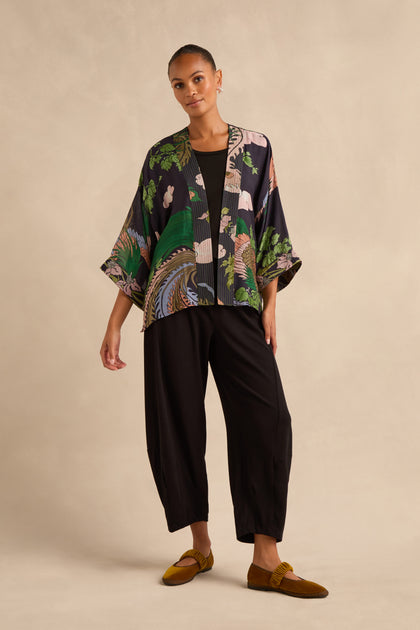 A person stands against a beige backdrop wearing the loose, patterned Phoenix Kimono, paired with black pants and mustard yellow loafers.