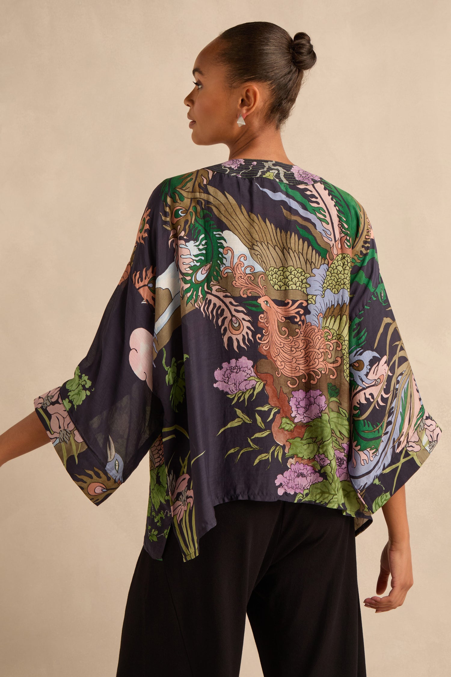 A woman is seen from behind wearing the Phoenix Kimono—a dark, loose modal blend piece adorned with colorful dragon and floral patterns—paired with black pants against a neutral background.