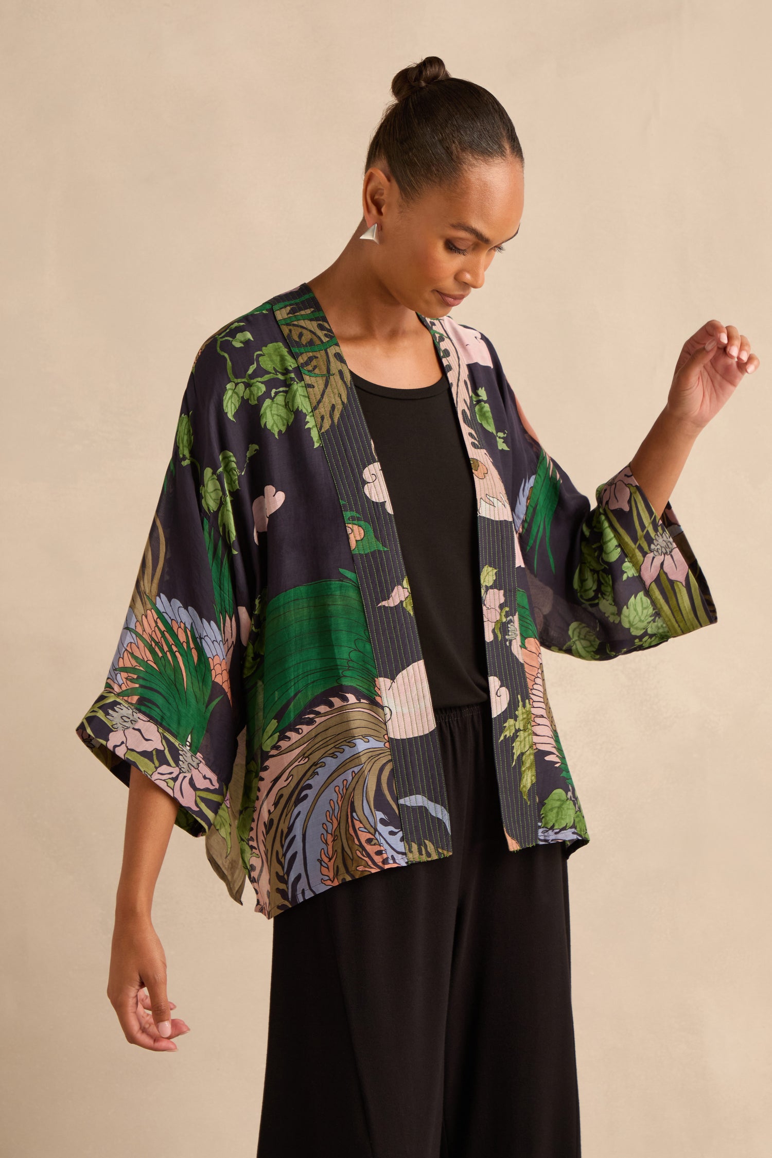 A woman wearing a black outfit with the vibrant Phoenix Kimono stands against a plain beige background, looking down with one arm slightly raised.