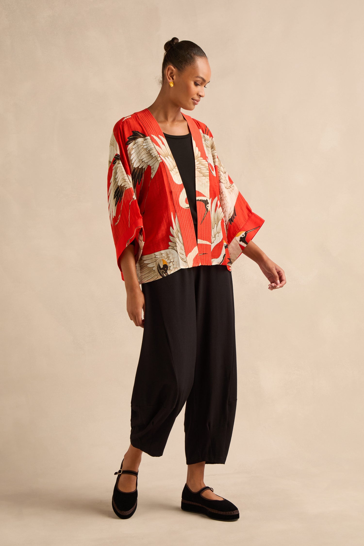 A woman stands against a neutral backdrop wearing the red Stork Kimono, featuring an Art Deco crane pattern, paired with a black top, loose black pants, and black shoes.