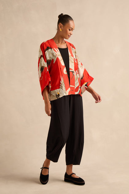 A woman stands against a neutral backdrop wearing the red Stork Kimono, featuring an Art Deco crane pattern, paired with a black top, loose black pants, and black shoes.