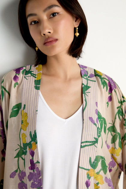 A woman wears the Wisteria Kimono, featuring a tan botanical print with purple and yellow flowers and green leaves, layered over a white V-neck shirt.
