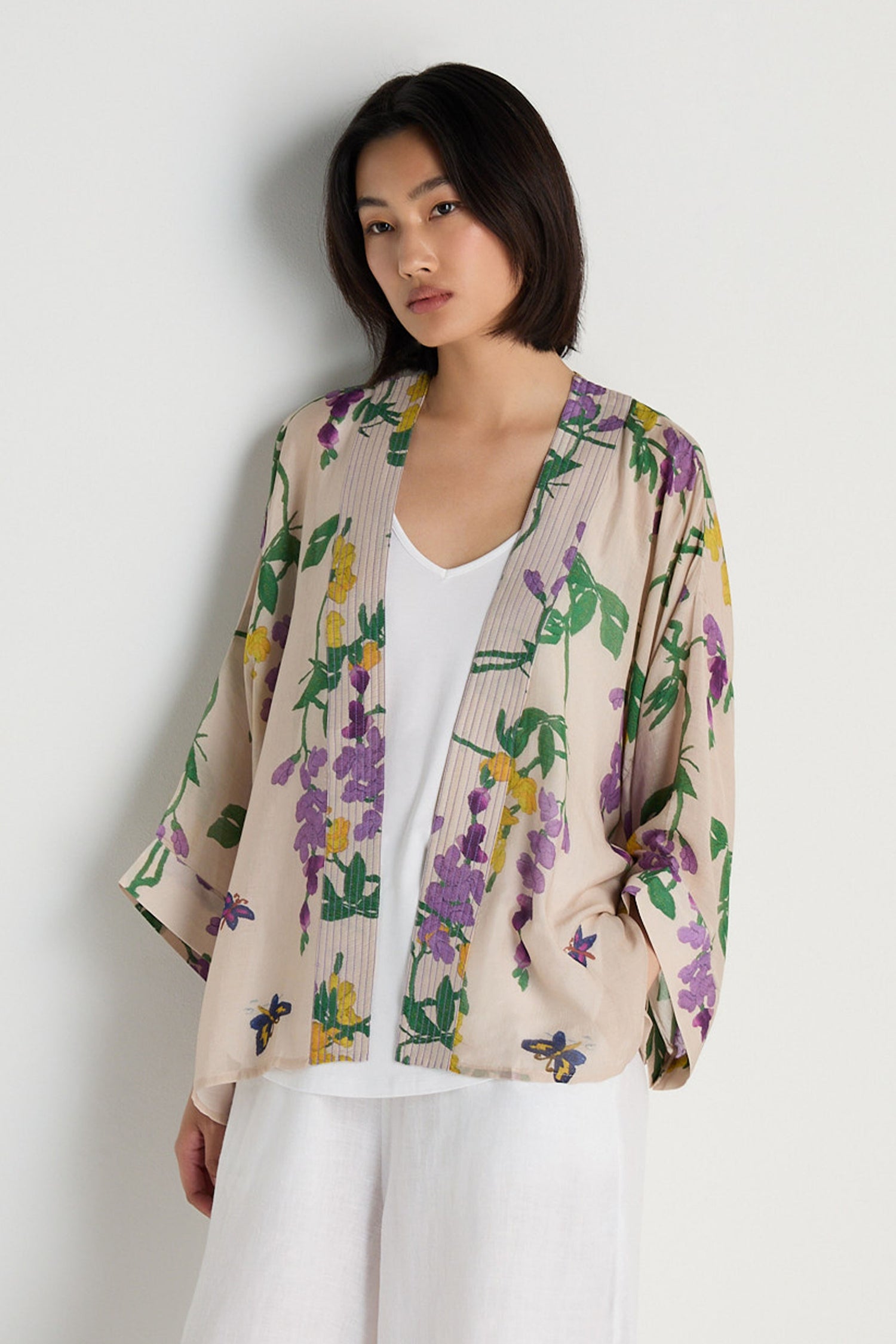 A woman models the Wisteria Kimono, featuring a cream base with purple and yellow floral patterns, layered over a white top and white pants against a plain white background.