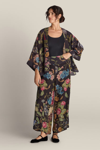 A person stands wearing a black floral kimono and matching Primula Palazzo Trouser over a black top. Their hair is styled back, and they are wearing black flat shoes. The background is plain beige.