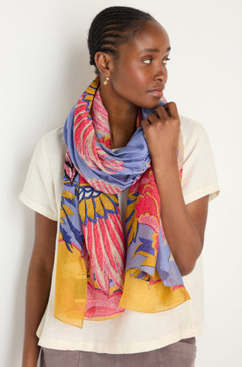 Woman wearing a cream short-sleeve top and the Crane Print Scarf—periwinkle with colorful floral accents, made from sustainable fabric—standing against a plain white background.