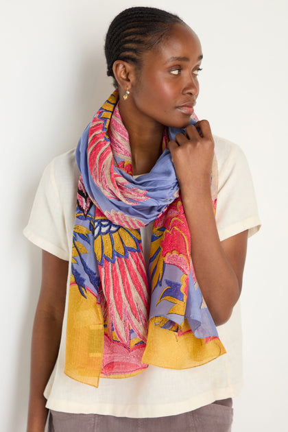 Woman wearing a cream short-sleeve top and the Crane Print Scarf—periwinkle with colorful floral accents, made from sustainable fabric—standing against a plain white background.