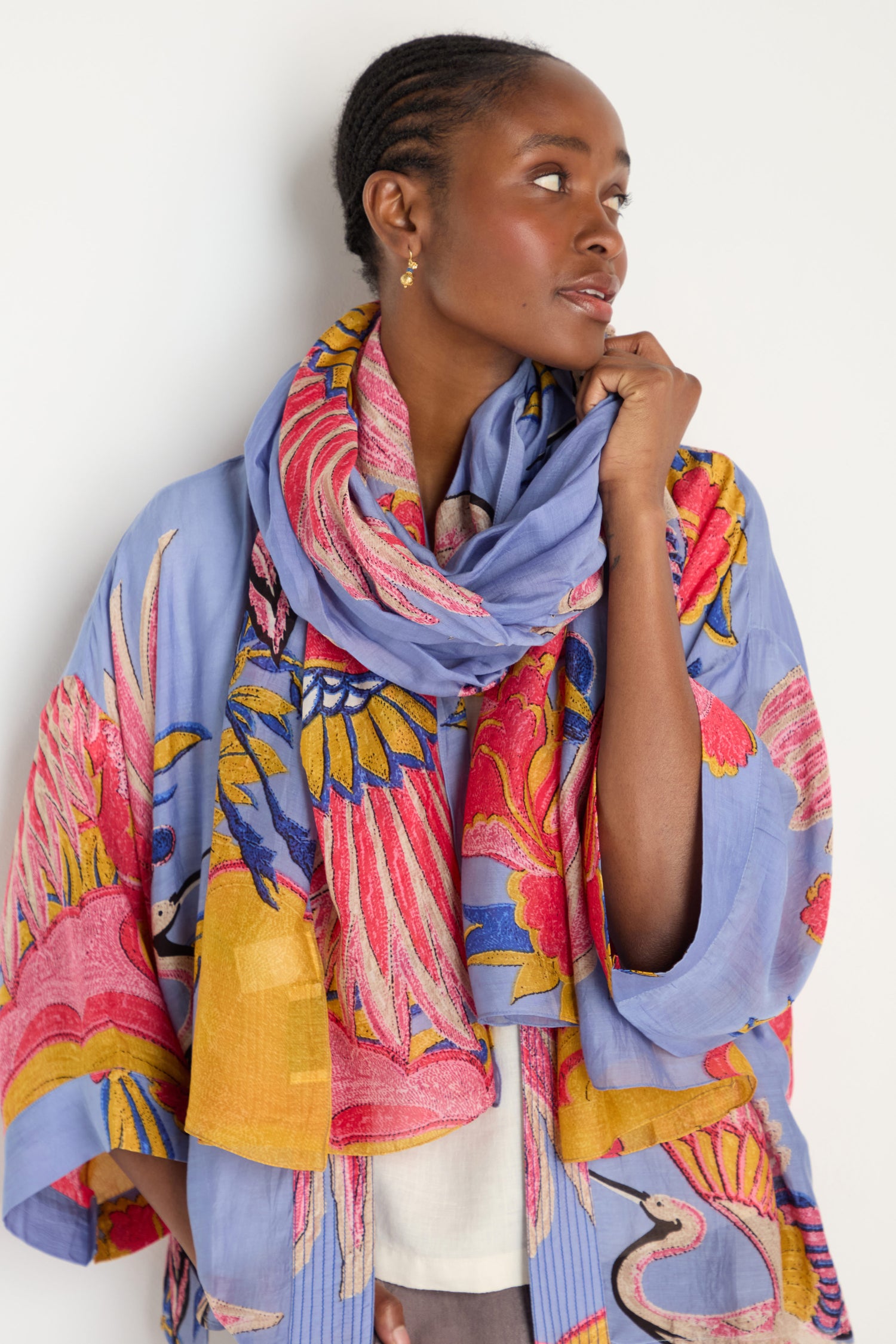 A woman stands against a white background, looking to the side, wearing the Crane Print Scarf—a periwinkle accessory featuring colorful floral and bird patterns—crafted from sustainable fabric.