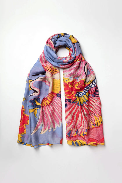 The Crane Print Scarf features a periwinkle base with colorful crane, flower, and leaf motifs in pink, yellow, red, and white. Made from sustainable fabric and shown flat on a white background.