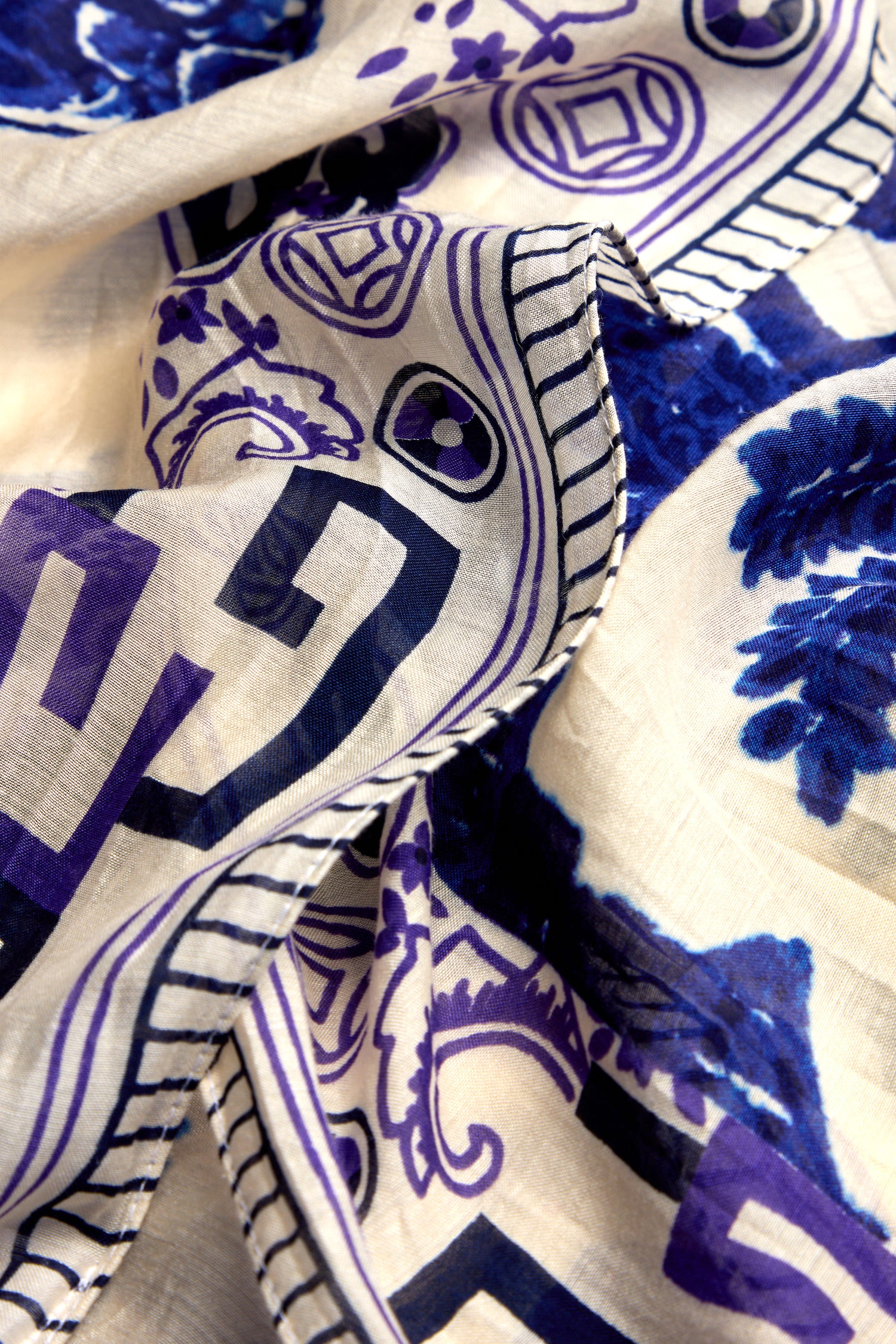 Close-up of a crumpled Willow Print Scarf, showcasing blue and purple floral patterns with geometric designs on a white background and stitched edges for timeless elegance.