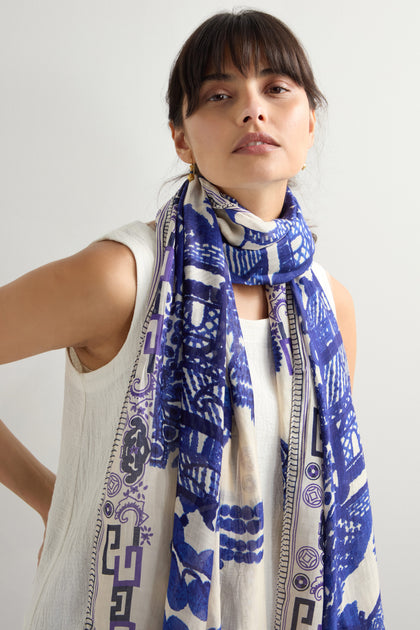 A person exudes timeless elegance as they look at the camera, wearing a white sleeveless top and a blue and white Willow Print Scarf.