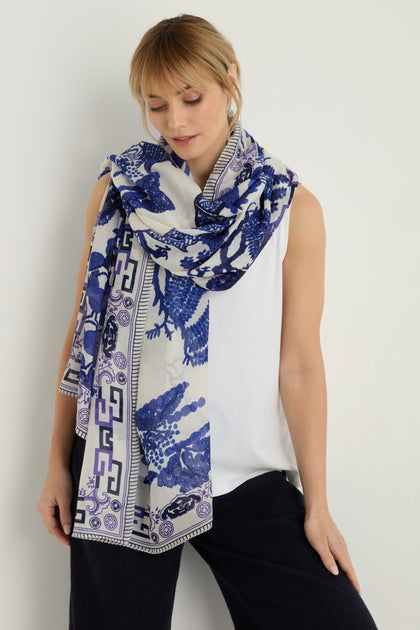 A woman stands against a plain light background, wearing black pants, a sleeveless white top, and our Willow Print Scarf—a large, lightweight scarf featuring blue patterned designs.