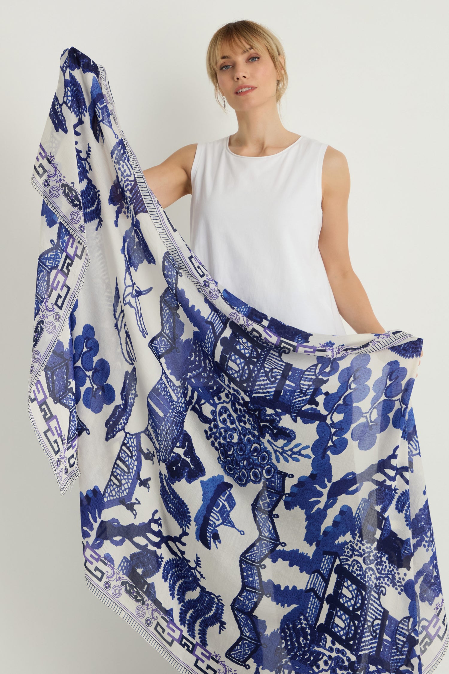 A woman in a white sleeveless top holds the Willow Print Scarf—a lightweight accessory with intricate blue and white designs—crafted from sustainable fabric, shown against a plain background.