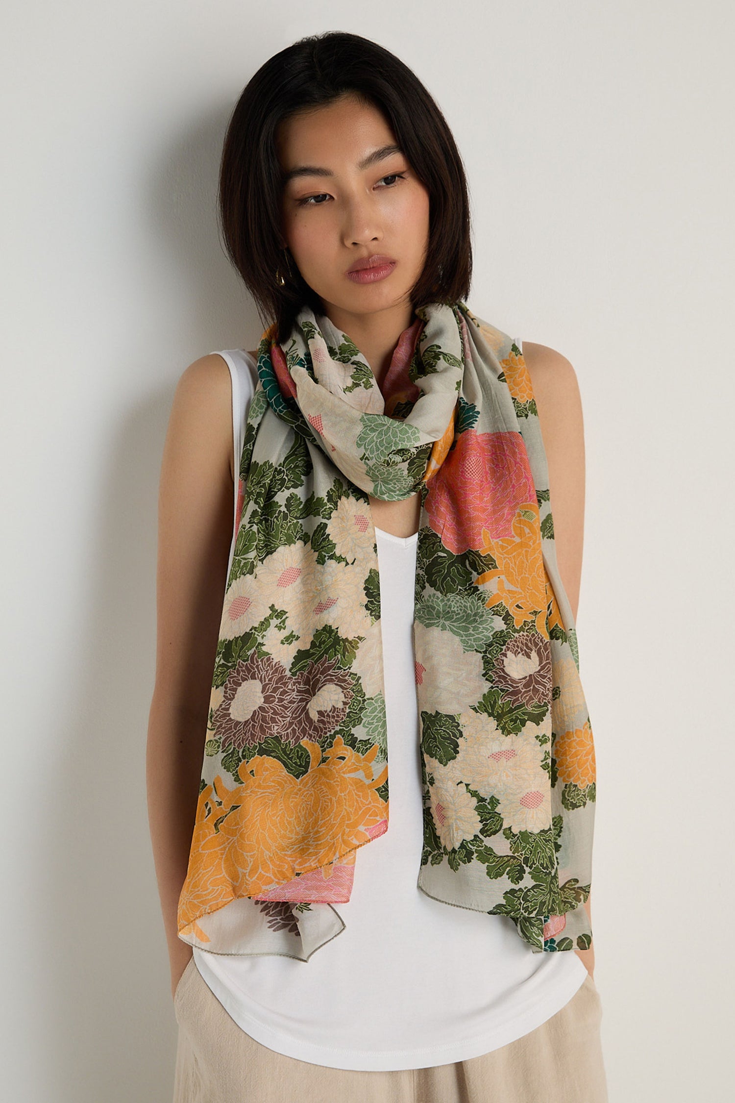 A woman stands against a plain light-colored wall, wearing a white sleeveless top and beige pants, styled with the Jacquard Scarf—a large, vintage-inspired floral accessory draped around her neck.