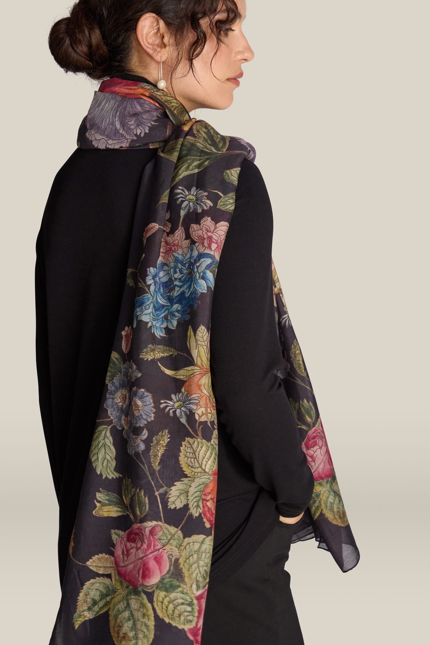 A woman wearing a black outfit and a long Primula Scarf draped over her shoulder is standing against a plain background, showcasing the elegance of sustainable fashion with this lovely floral print accessory.