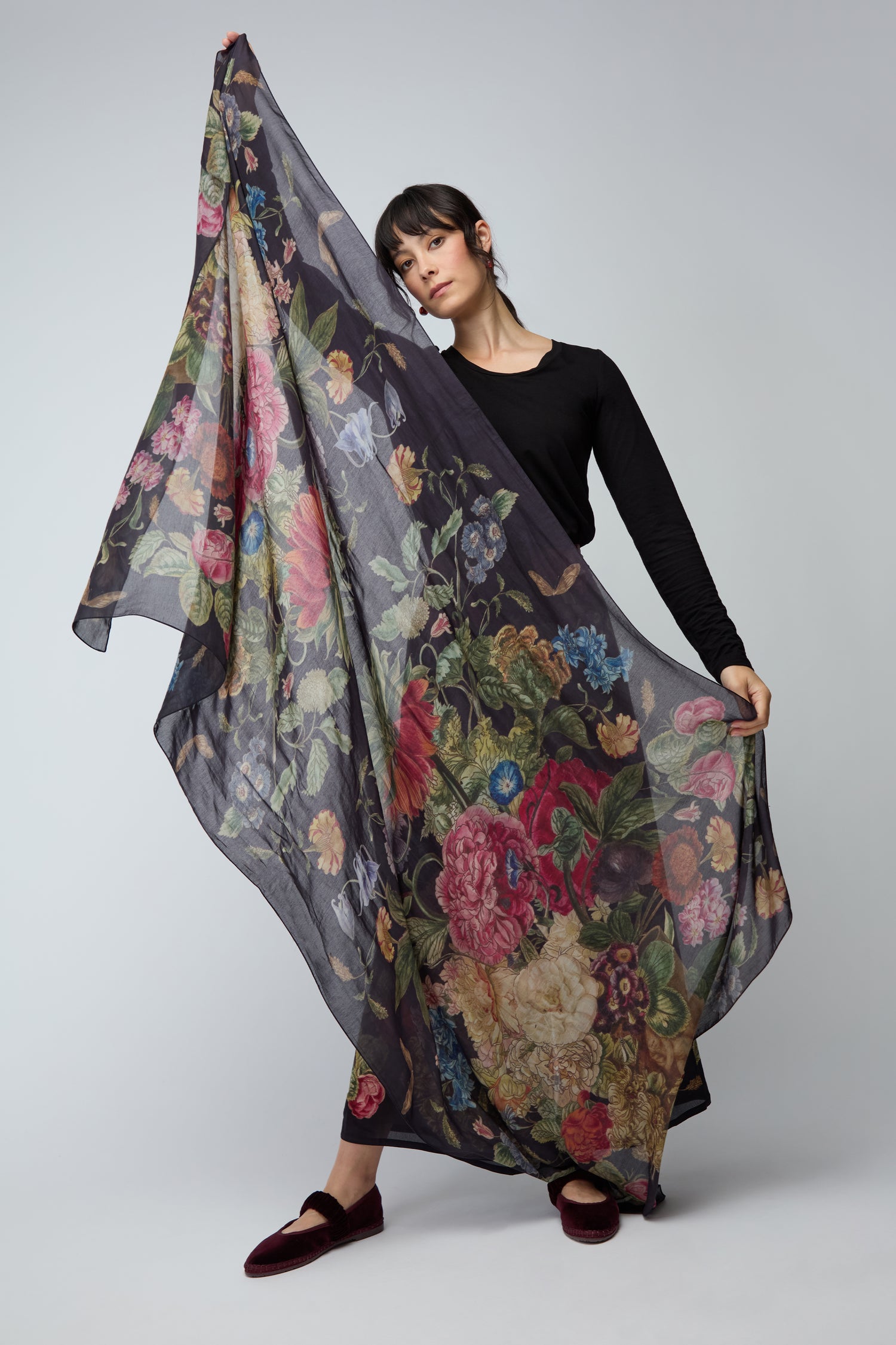 A person in black displays the Primula Scarf, a large, sheer crepe fabric accessory featuring a vibrant floral pattern, set against a plain background.