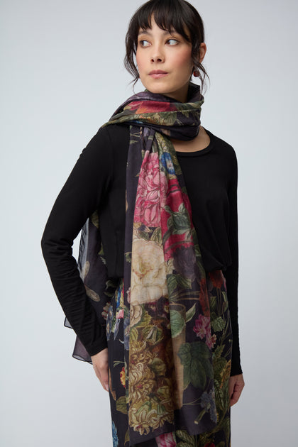 A woman stands against a plain light gray background, wearing a black long-sleeve top and the Primula Scarf, a floral accessory draped elegantly over her shoulders.
