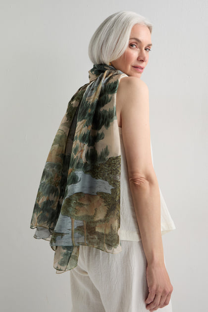 A woman with short gray hair wears a sleeveless white top and pants, elegantly draping a sheer Rivers and Trees Print Scarf—nature-inspired with a landscape design—over her shoulders.