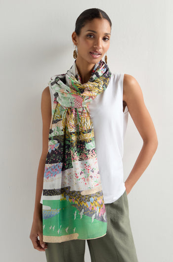 A woman stands against a white background wearing a white sleeveless top, green pants, and a Spring Print Scarf with oriental garden silhouettes. She accessorizes with gold earrings and has her hair pulled back.