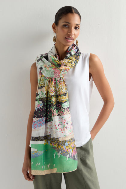 A woman stands against a white background wearing a white sleeveless top, green pants, and a Spring Print Scarf with oriental garden silhouettes. She accessorizes with gold earrings and has her hair pulled back.