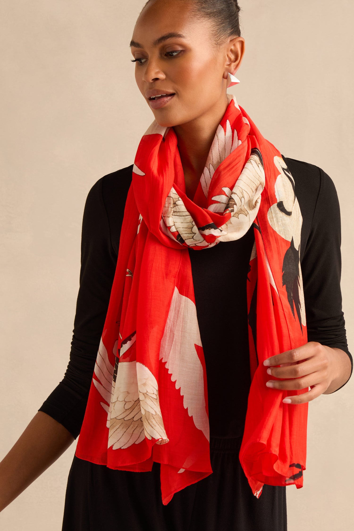 A woman wearing a black top and the Stork Scarf, featuring a bright red background with a white and beige bird wing pattern, stands against a neutral backdrop.