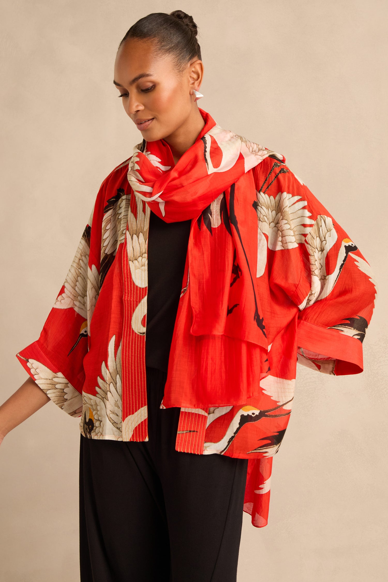 A woman stands against a beige background, wearing a red patterned kimono-style jacket and the Stork Scarf featuring white cranes, draped over a black outfit.