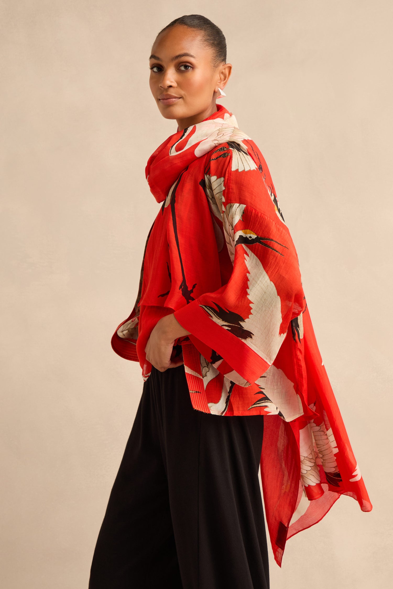 A woman stands against a beige background, wearing a red floral kimono-style top and black pants, her look elegantly finished with the Stork Scarf draped loosely around her neck as she looks at the camera.
