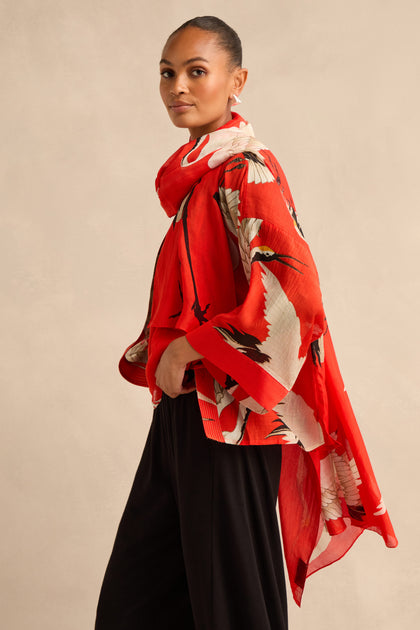 A woman stands against a beige background, wearing a red floral kimono-style top and black pants, her look elegantly finished with the Stork Scarf draped loosely around her neck as she looks at the camera.