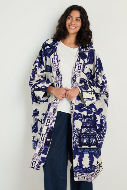 A person wearing a white shirt, blue jeans, and the Willow Print Summer Coat—a blue and white patterned summer layer—stands smiling against a plain light background.