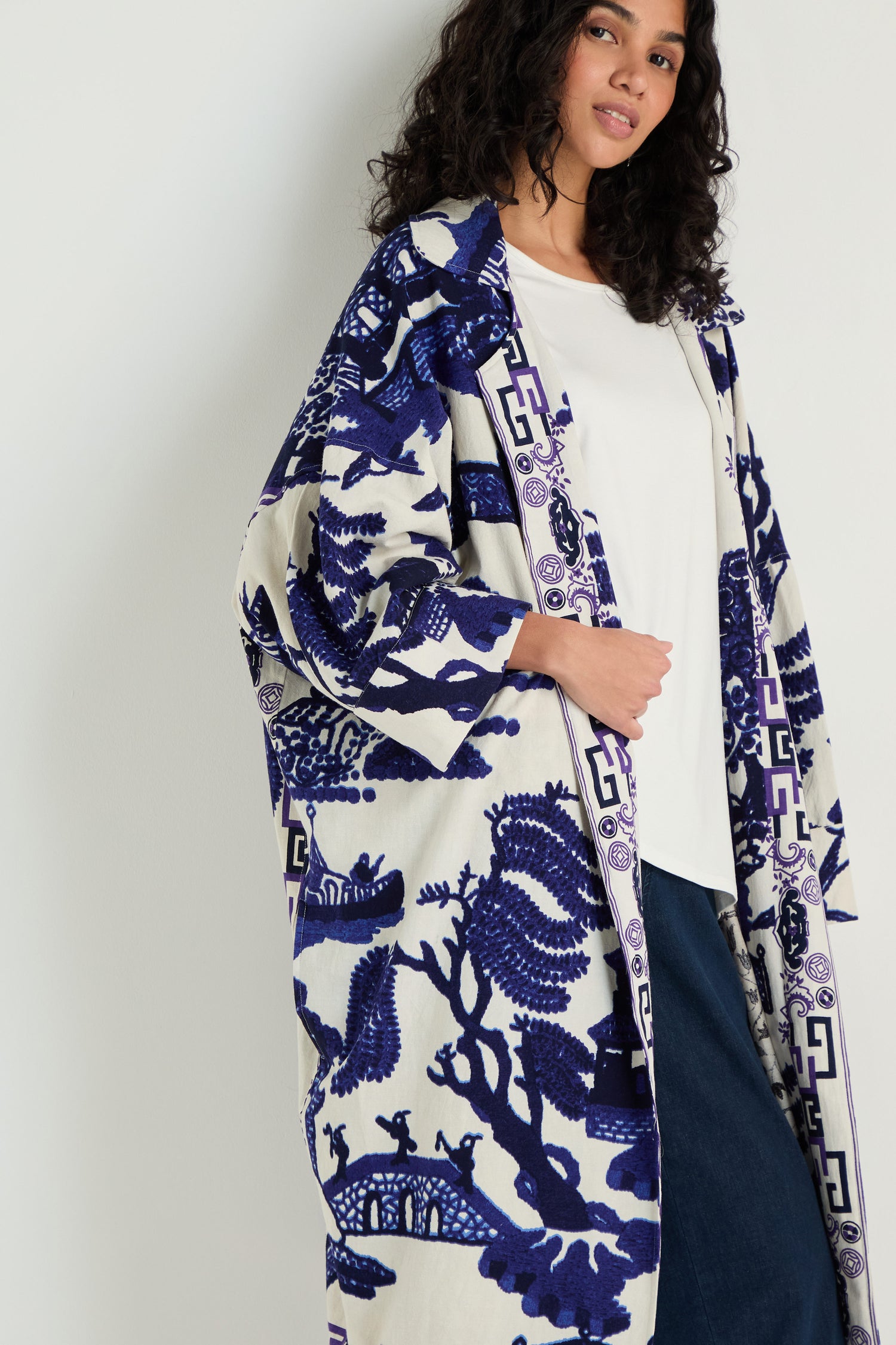 A woman in a white shirt and blue jeans stands by a wall, draped in the Willow Print Summer Coat—a lightweight layer featuring elegant blue and white patterns.