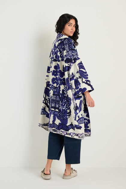 A person with dark wavy hair wears the Willow Print Summer Coat, featuring blue and white chinoiserie patterns, paired with wide-leg jeans and beige shoes, standing against a plain white wall.