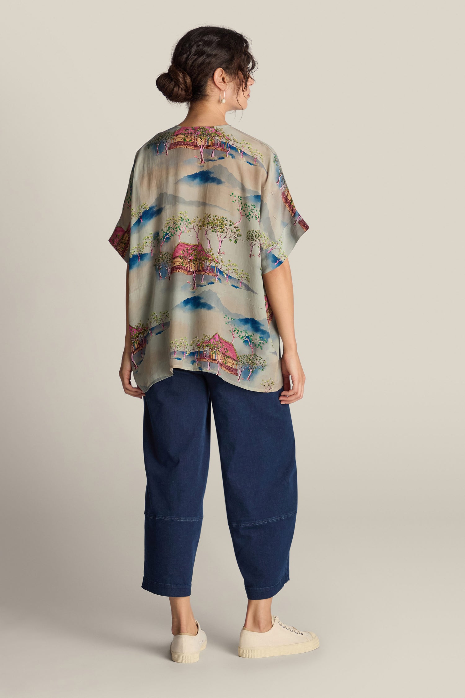 A person stands with their back to the camera, sporting a loose-fitting Landscape Blouse adorned with wearable works of art—colorful prints reminiscent of Japanese landscape watercolors—and blue cropped pants, paired with light-colored shoes.