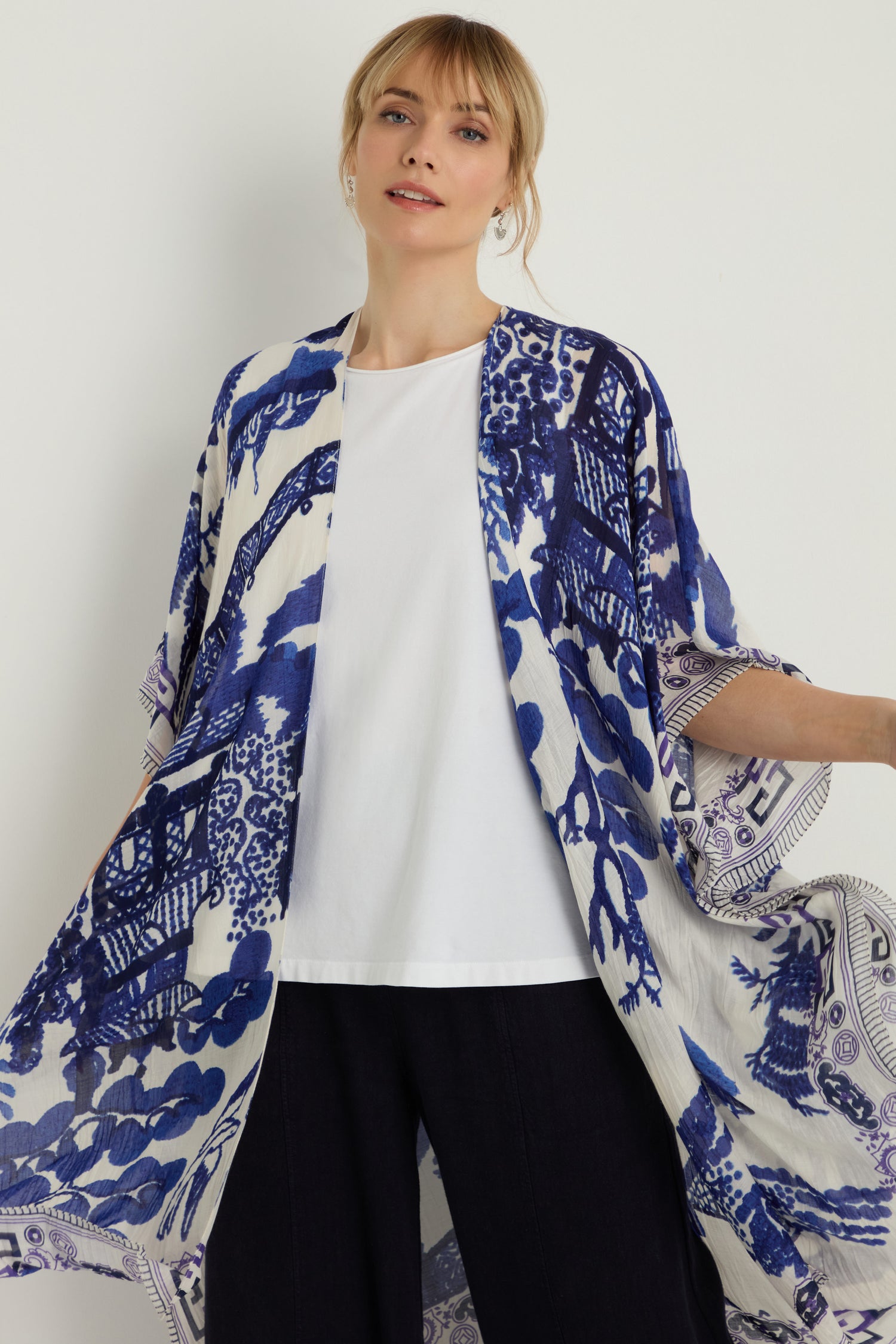 A woman stands against a plain background, wearing a white t-shirt, dark pants, and the Willow Print Throwover—a blue-and-white kimono-style jacket with wide sleeves that offers a stylish and versatile layering option.