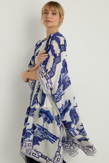A woman models the Willow Print Throwover, a white and blue kimono-style cover-up featuring animal and geometric prints on sustainable fabric. She stands against a light background, highlighting this versatile garment.