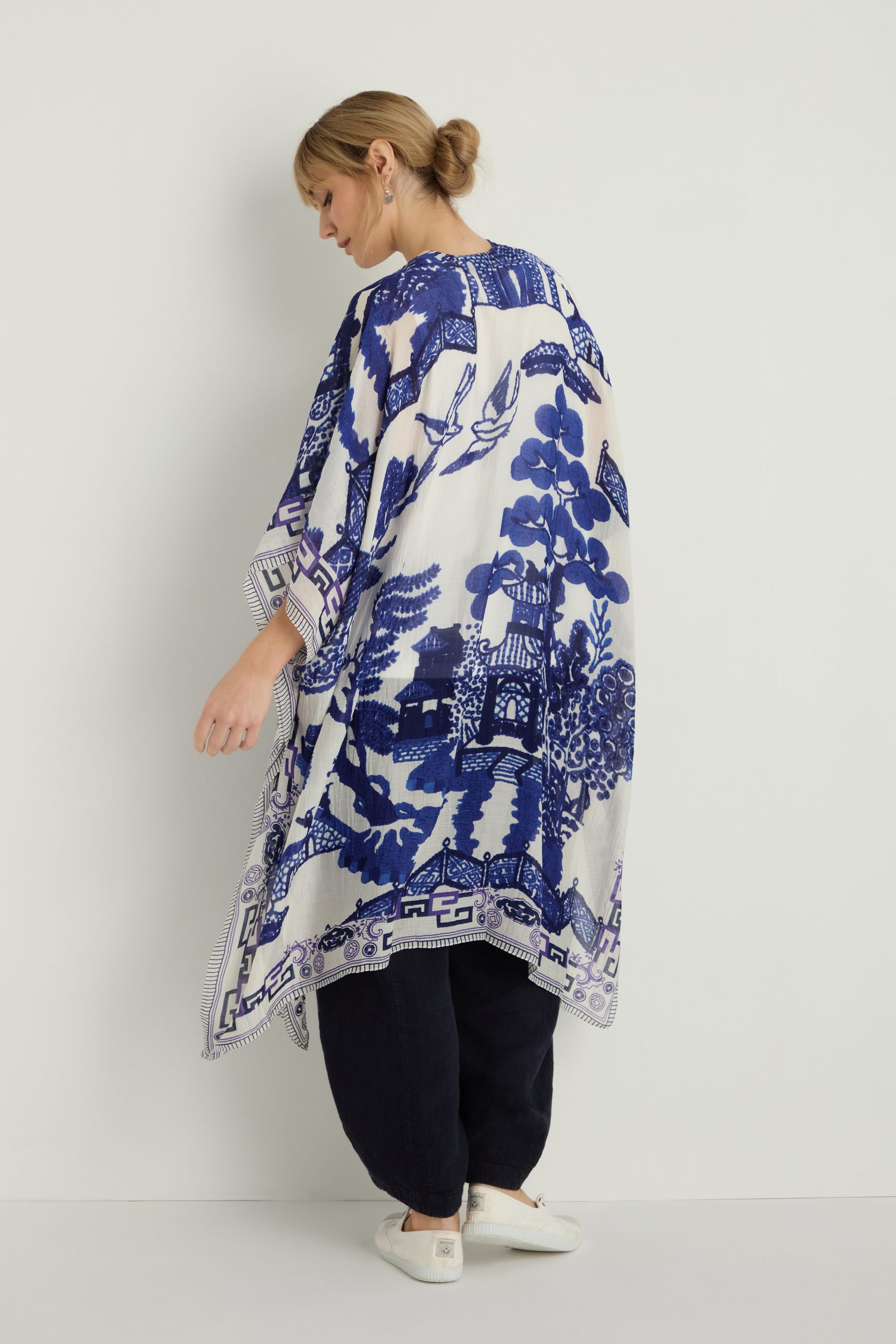 A woman, facing away from the camera, wears the Willow Print Throwover—made from sustainable fabric—against a white wall, paired with black pants and white shoes.