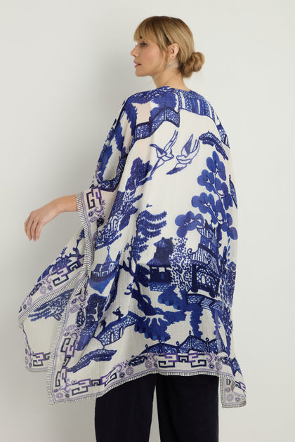 A woman with her back turned wears the Willow Print Throwover, a loose white and blue kimono made from sustainable fabric, adorned with an intricate scenic print featuring birds, trees, and classic architecture.
