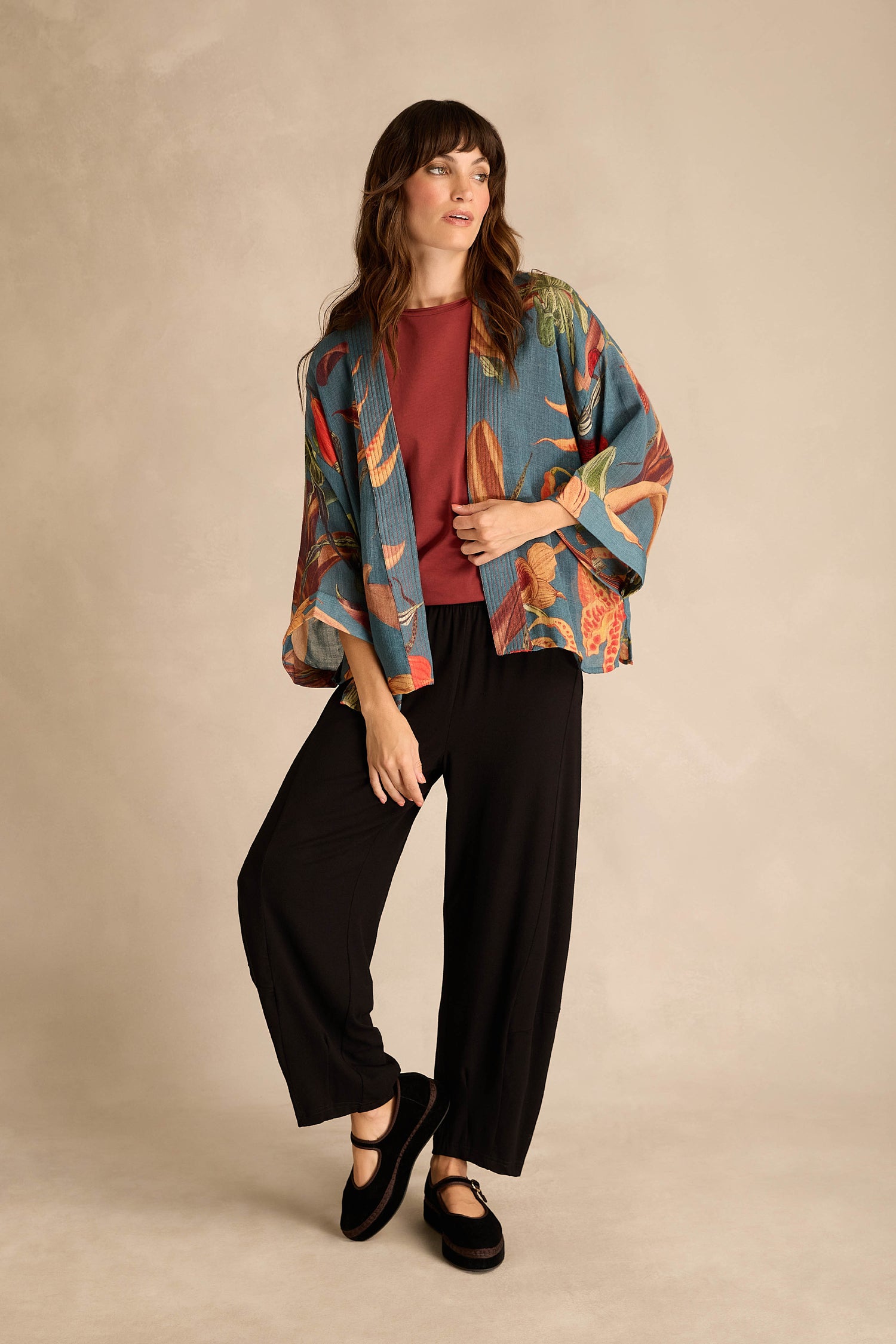 A woman stands against a neutral backdrop wearing the Orchid Wool Kimono, featuring an oversized slipper orchid print, layered over a rust top, black wide-leg pants, and black shoes—an elegant winter layer.