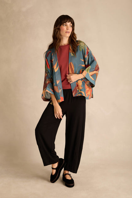 A woman stands against a neutral backdrop wearing the Orchid Wool Kimono, featuring an oversized slipper orchid print, layered over a rust top, black wide-leg pants, and black shoes—an elegant winter layer.