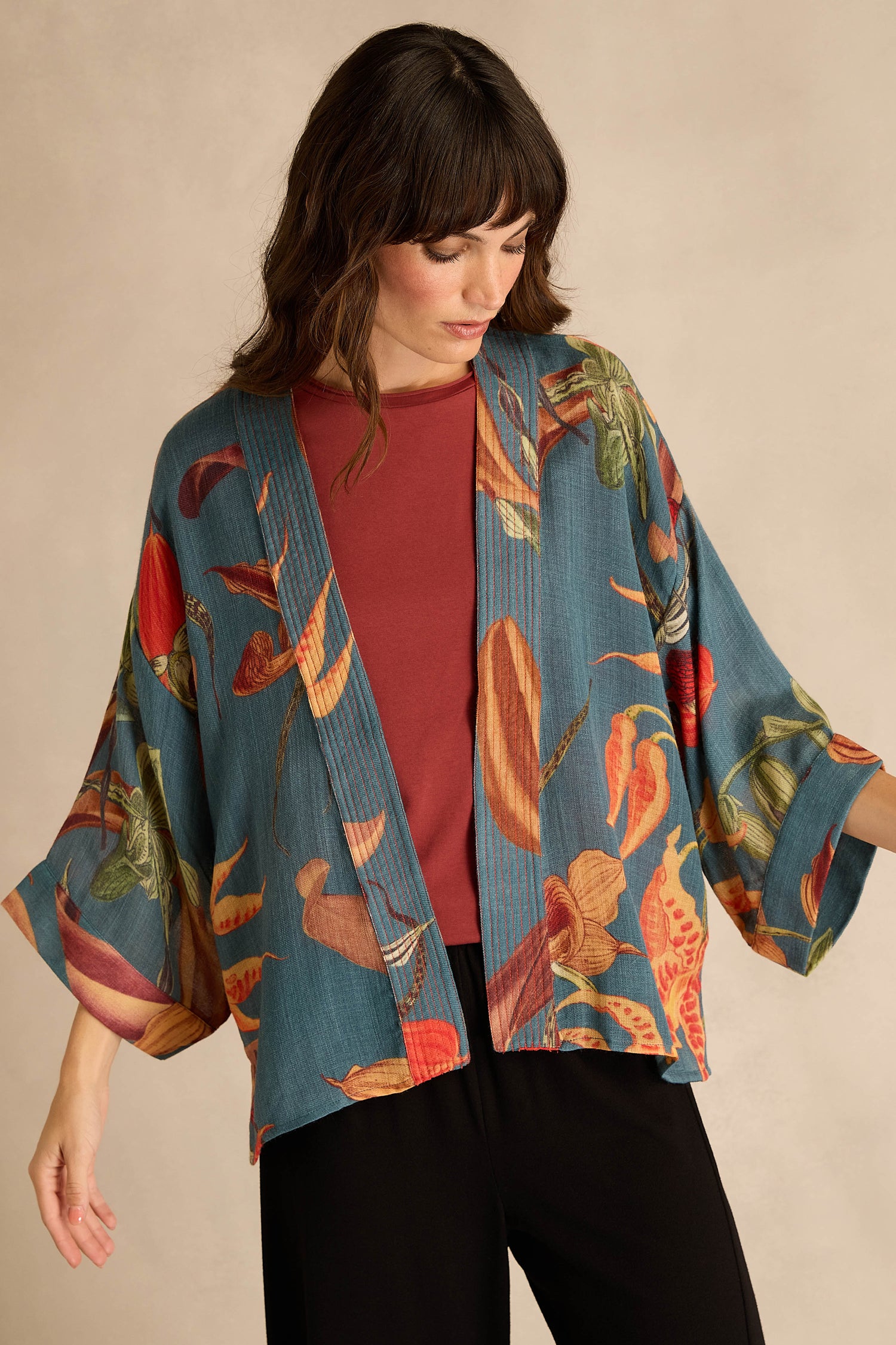 Model wears the Orchid Wool Kimono—a blue kimono-style jacket with an oversized slipper orchid print—paired with a red shirt and black pants, standing against a neutral background; perfect as a vibrant winter layer.