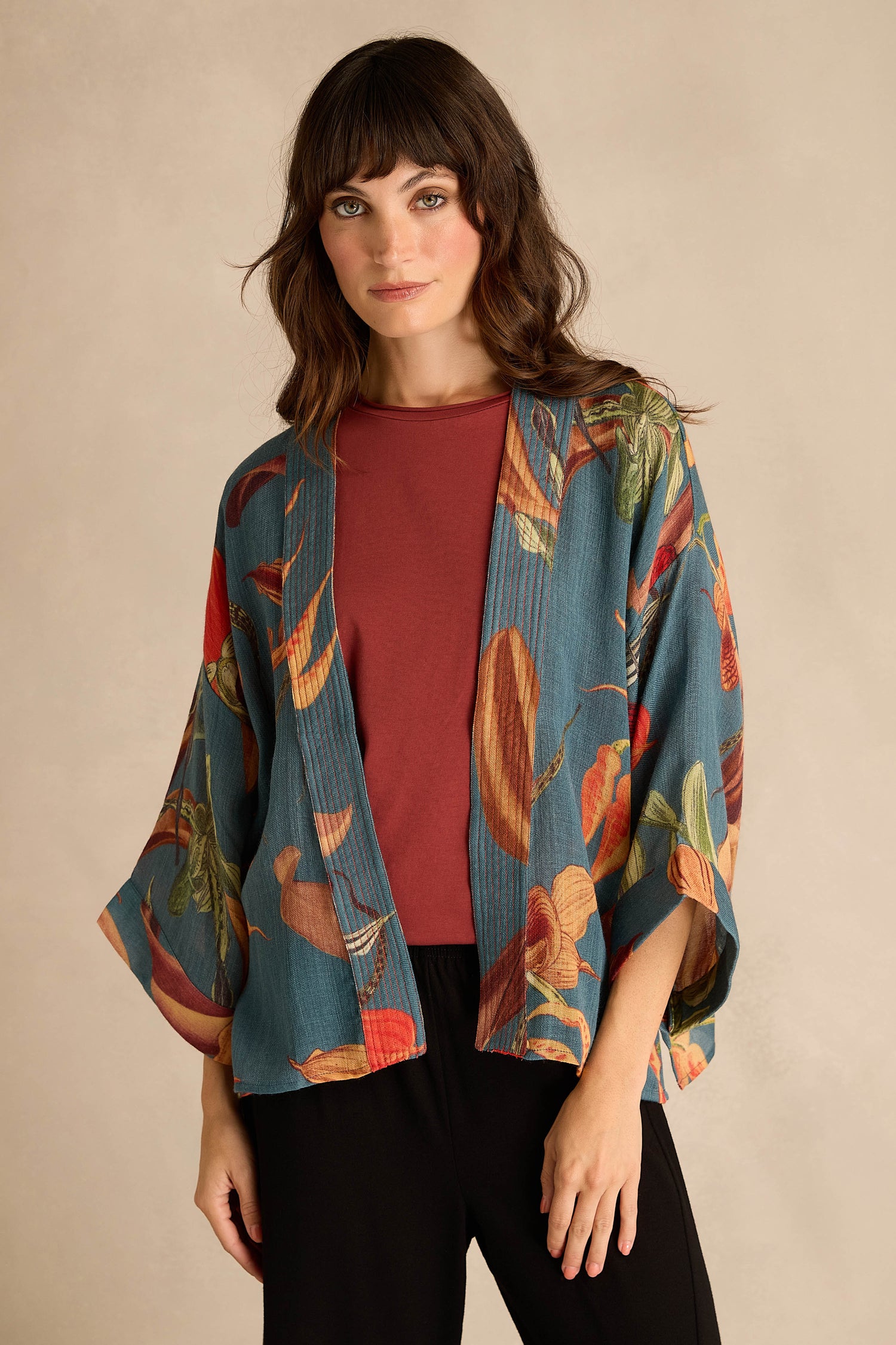 A woman models the Orchid Wool Kimono, adorned with an oversized slipper orchid print, layered over a rust-colored top and black pants against a plain beige backdrop—an elegant winter wardrobe addition.