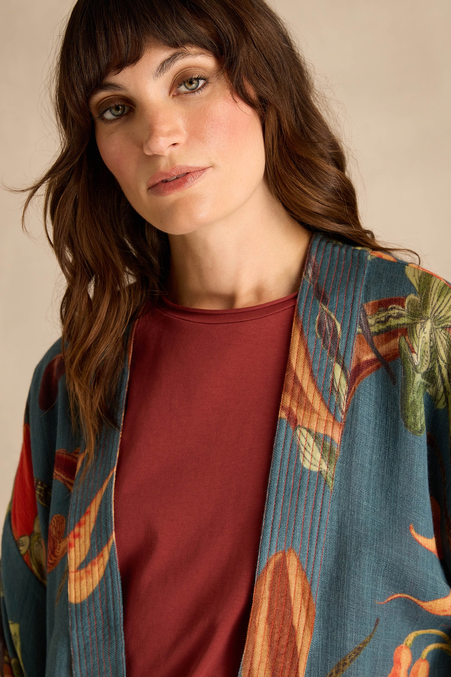 A woman with wavy brown hair wears a red top and the Orchid Wool Kimono, which showcases an oversized slipper orchid print, as she gazes slightly to the side against a neutral background—a perfect winter layer that adds bold floral elegance.