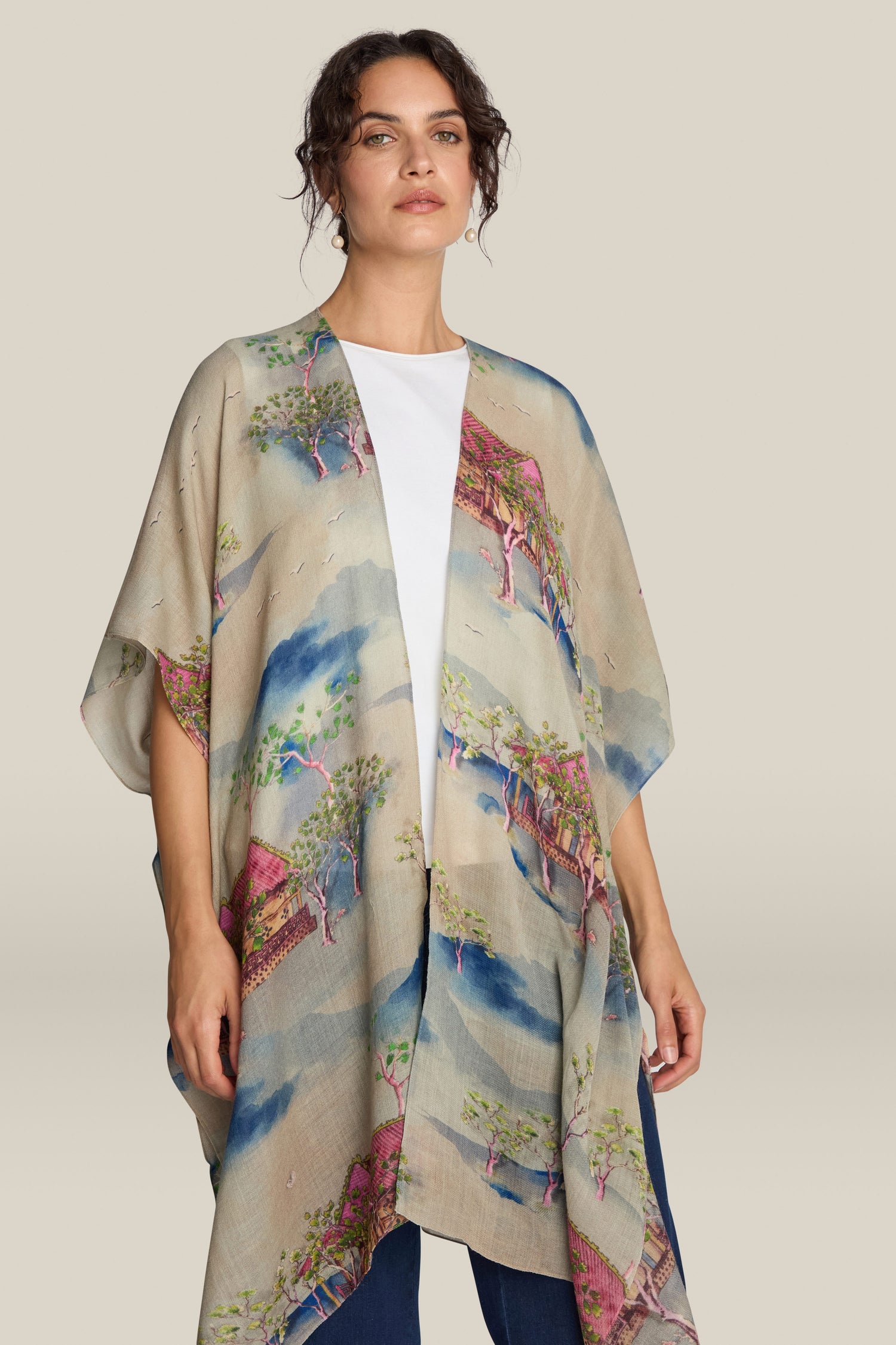 A person stands wearing the Landscape Wool Throwover, a light beige kimono with unique prints reminiscent of Japanese watercolours, over a white shirt and blue jeans, set against a neutral background.