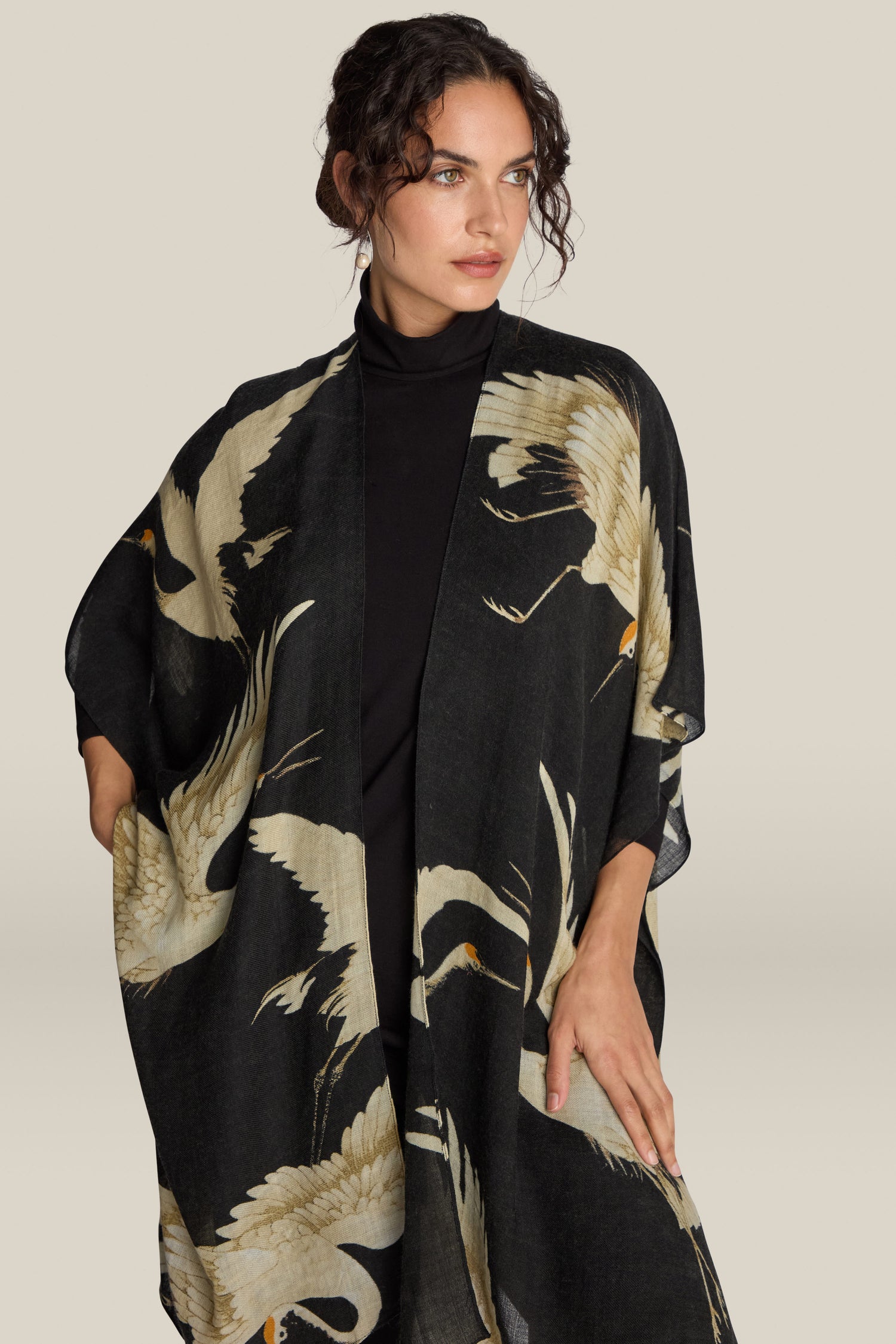 A woman models the sophisticated Stork Wool Throwover from One Hundred Stars, an elegant black kimono-style shawl adorned with a striking crane bird pattern.