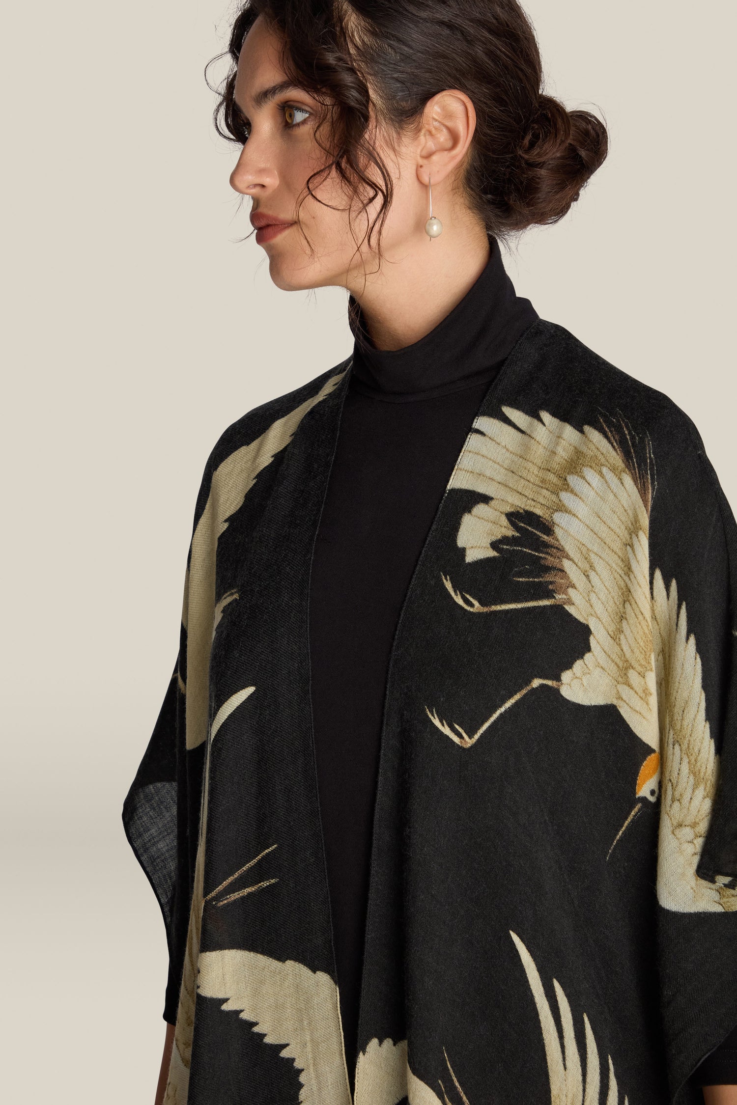 A woman with dark hair tied back is wearing a black turtleneck and a Stork Wool Throwover featuring a crane design. She is viewed from a slight side angle against a plain background.