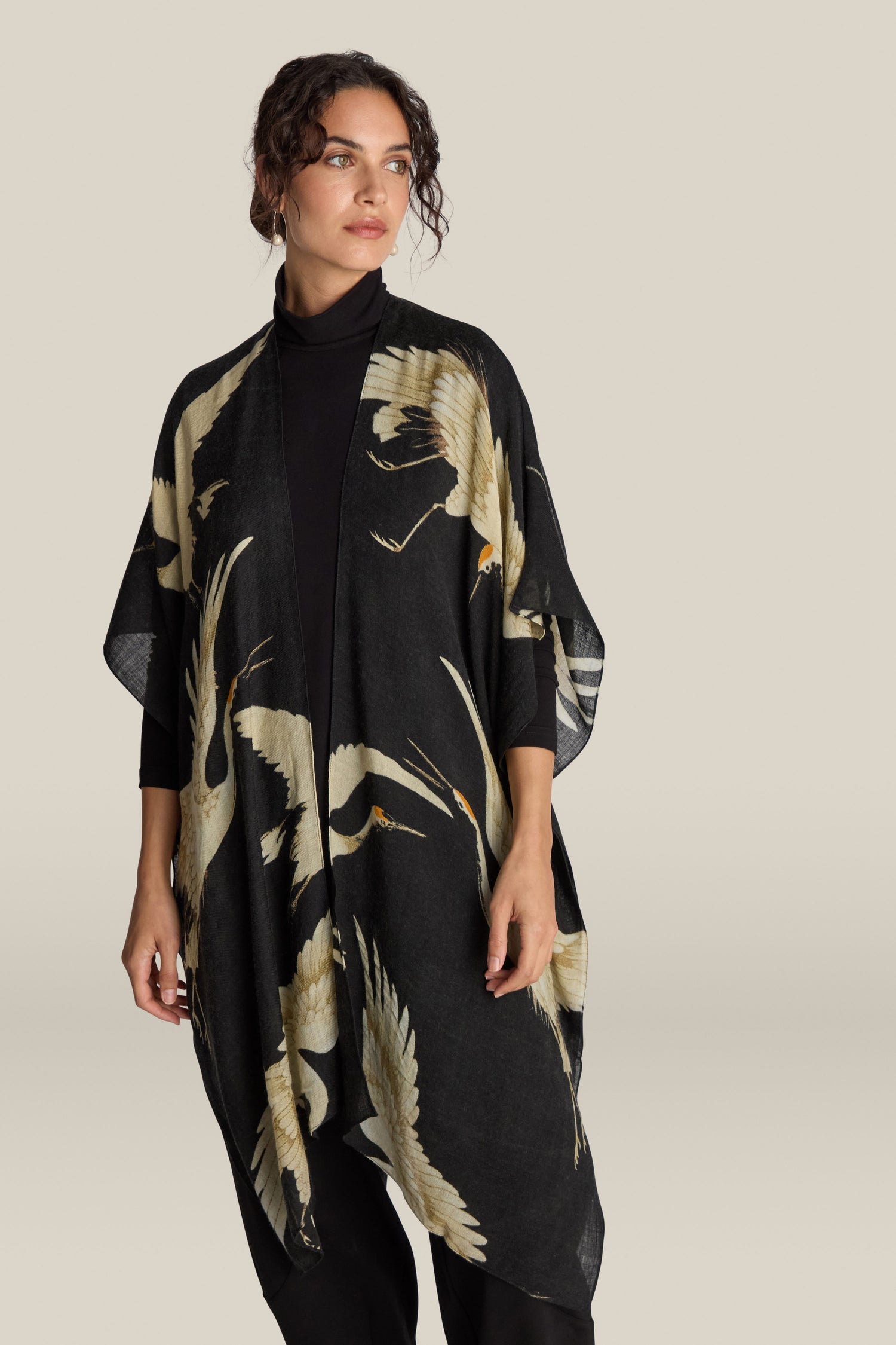 A woman wearing a black kimono-style wrap with a crane bird pattern stands against a beige background, elegantly accessorizing her look with the luxurious Stork Wool Throwover.