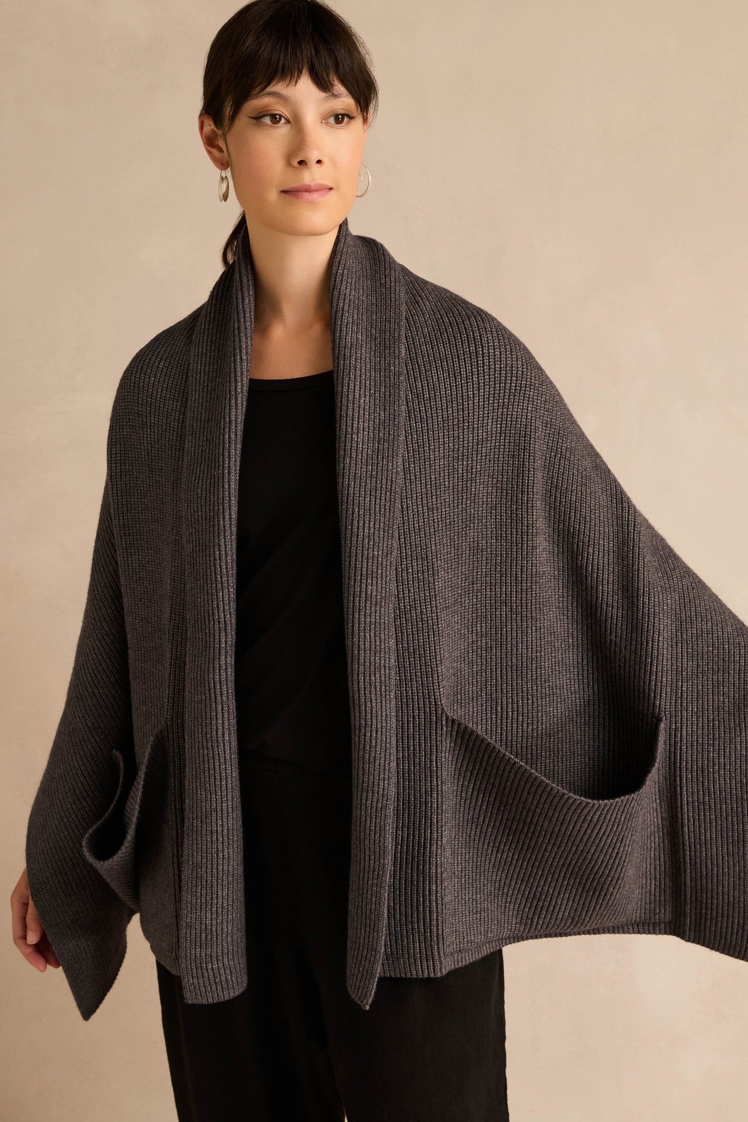 Person wearing the Charcoal Pocket Scarf, a dark gray accessory with oversized pockets and a cozy ribbed knit texture, styled over a black shirt and black pants against a neutral background. Crafted from a recycled fibre blend for added sustainability.