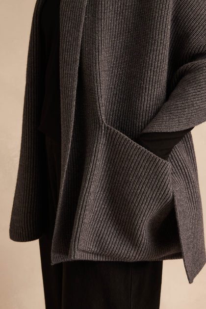 A person with one hand in the oversized pocket of the Charcoal Pocket Scarf stands against a neutral background. Only the torso and part of the arm are visible, drawing attention to the cozy design crafted from a recycled fibre blend.