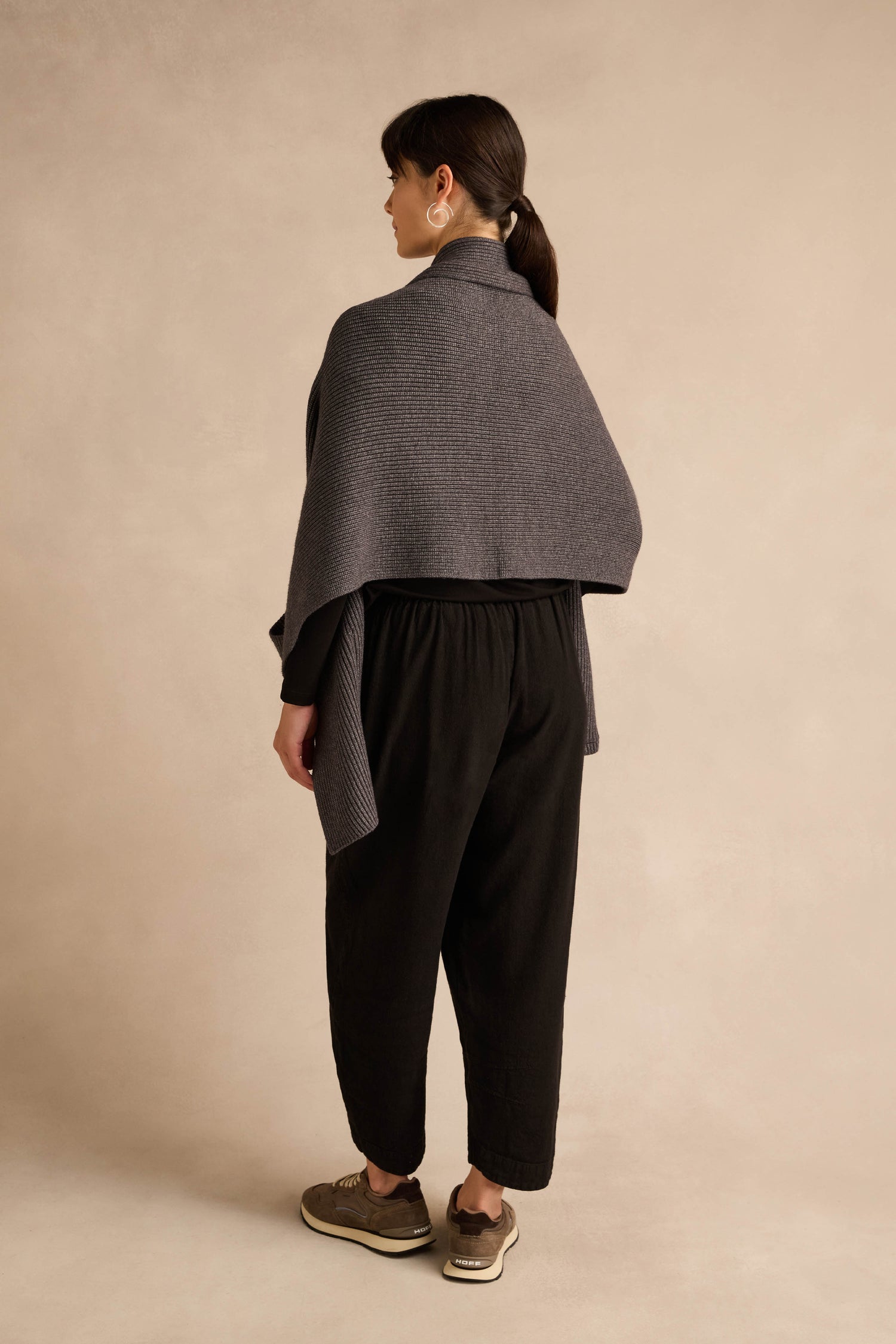 A woman stands with her back to the camera, wearing the Charcoal Pocket Scarf with oversized pockets, paired with black pants and sneakers, against a plain beige background.