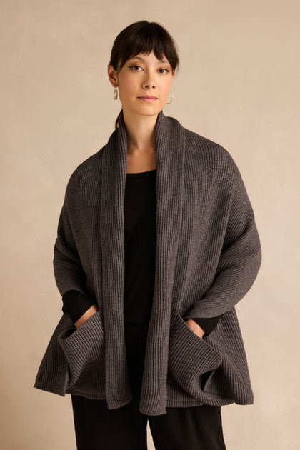 A woman stands facing forward, draped in the Charcoal Pocket Scarf crafted from a recycled fibre blend, layered over a black outfit. Her hands rest in the scarf’s oversized pockets, and she is set against a neutral beige background.